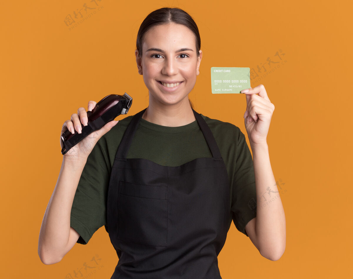 微笑微笑着的年轻理发女孩穿着制服拿着理发器和信用卡在橙色理发师头发剪子
