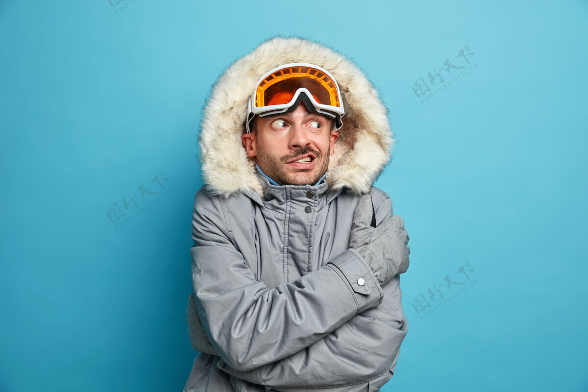 人不满的未刮胡子的年轻人因寒冷而颤抖 他戴着滑雪镜和冬衣拥抱着自己成人快乐摇晃