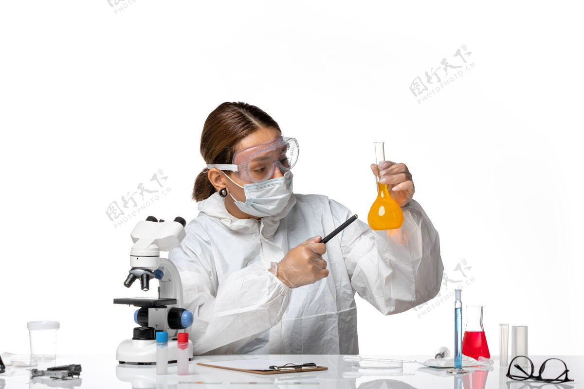 专业人员正面图女医生穿着特制的工作服 戴着口罩 拿着装有黄色药瓶的白色办公桌上的大流行冠状病毒口罩指向视图