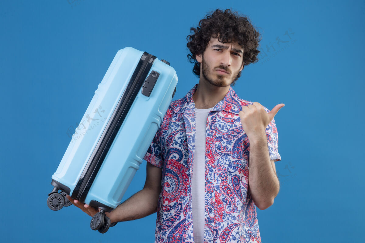 困惑迷茫的年轻英俊的旅行家男子拿着手提箱指着右边的蓝色空间和复印空间指向手提箱帅哥