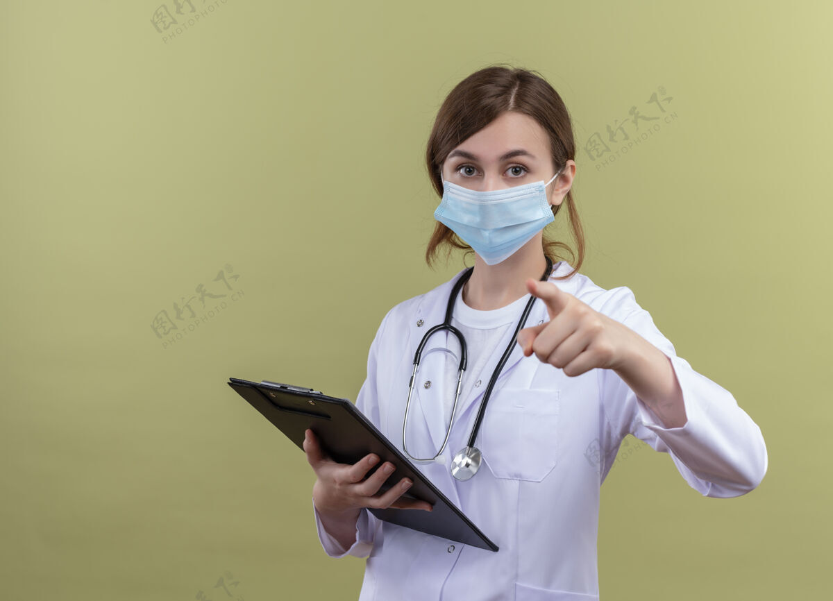 信心自信的年轻女医生穿着医用长袍 戴着口罩 手持听诊器 拿着剪贴板 指着与世隔绝的绿色空间和复印空间听诊器面具持有