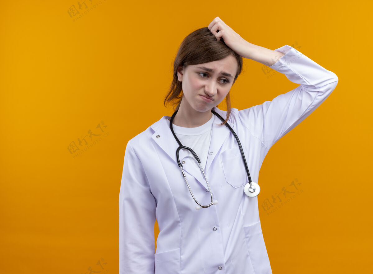 手焦急的年轻女医生穿着医用长袍 戴着听诊器 将手放在头上 放在隔离的橙色空间和复印空间穿听诊器医学