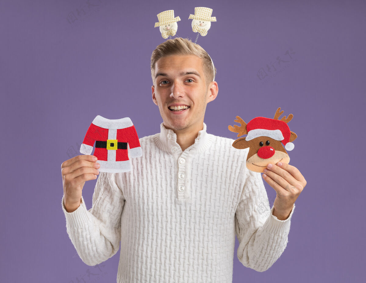 雪人快乐的年轻帅哥戴着雪人的头带 拿着圣诞老人的外套和驯鹿的纸饰品 看着孤立在紫色背景上的相机圣诞老人小伙子紫色