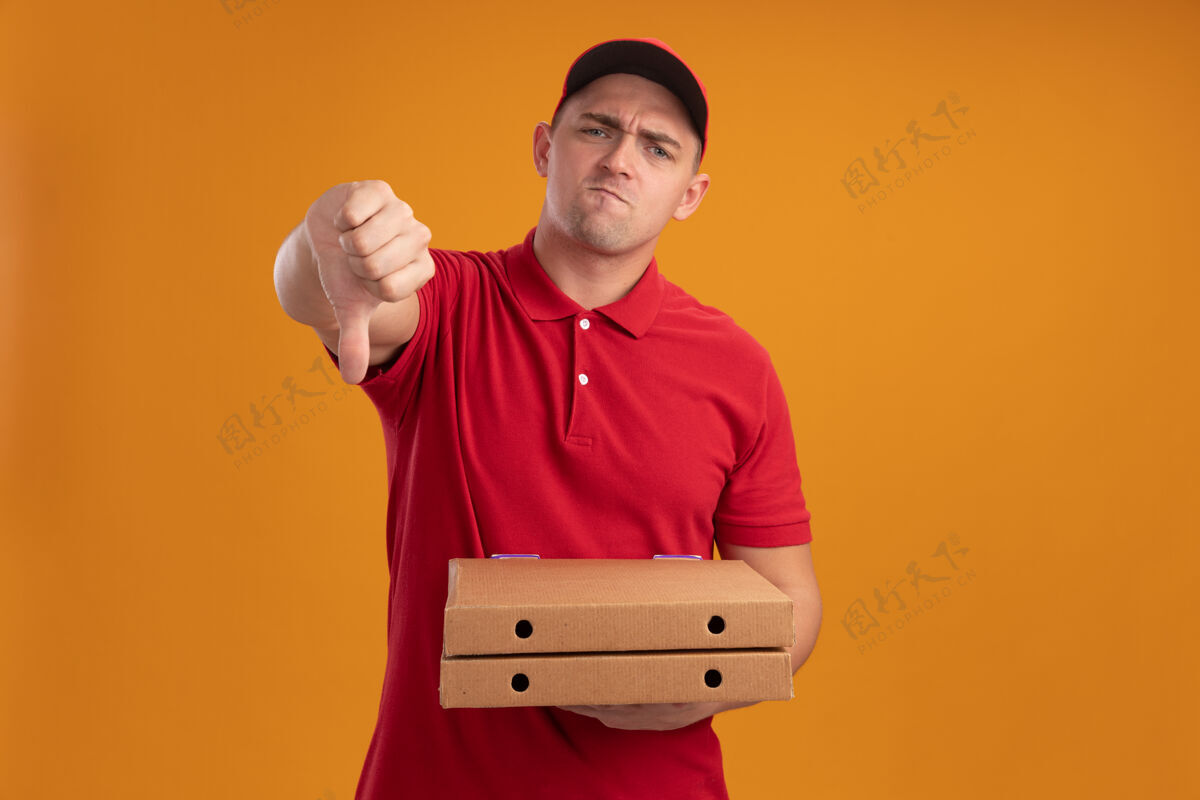 放下年轻的送货员穿着制服 戴着帽子 拿着披萨盒 拇指朝下 被隔离在橙色的墙上不愉快公民人