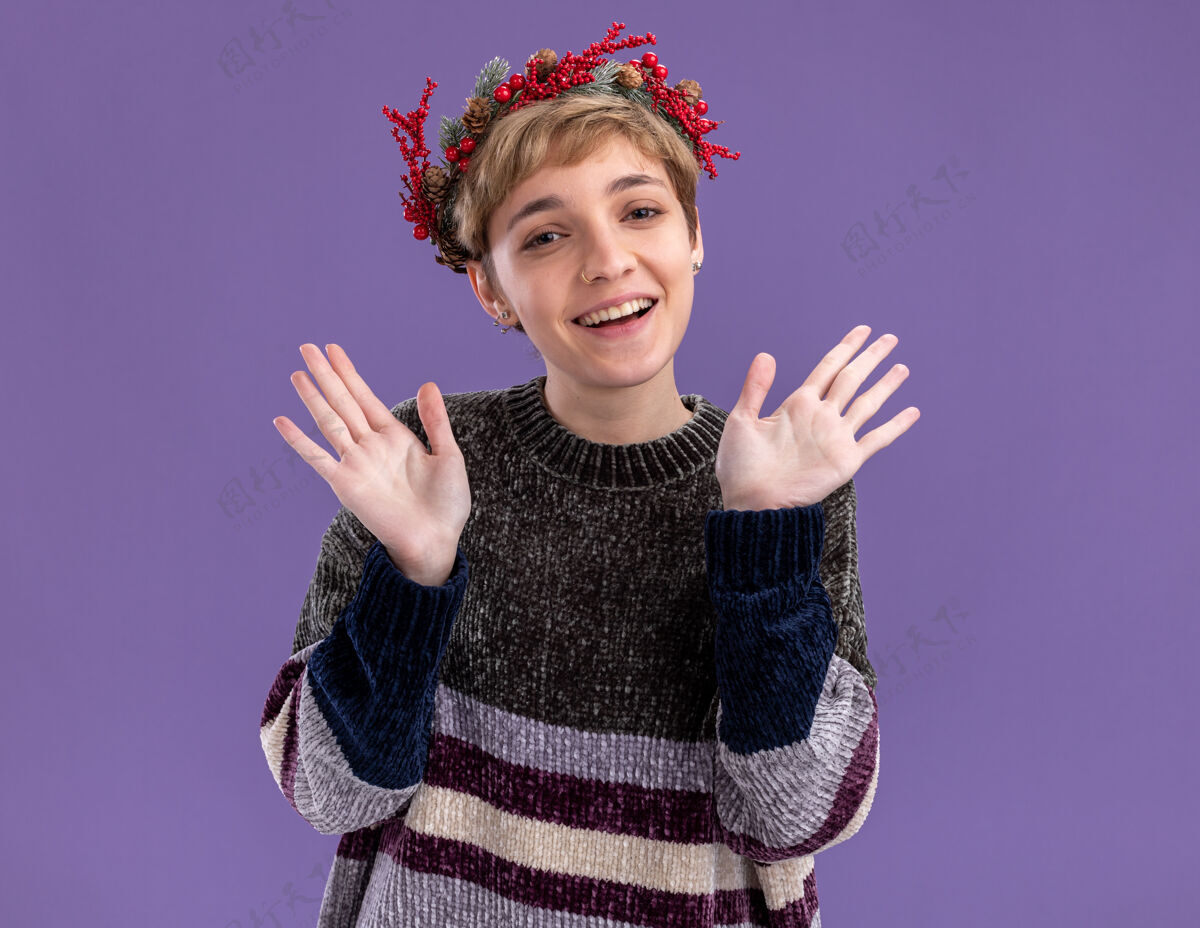 手快乐的年轻漂亮女孩戴着圣诞花环看着镜头 在紫色的背景下 两只空荡荡的手被孤立了表演圣诞快乐圣诞