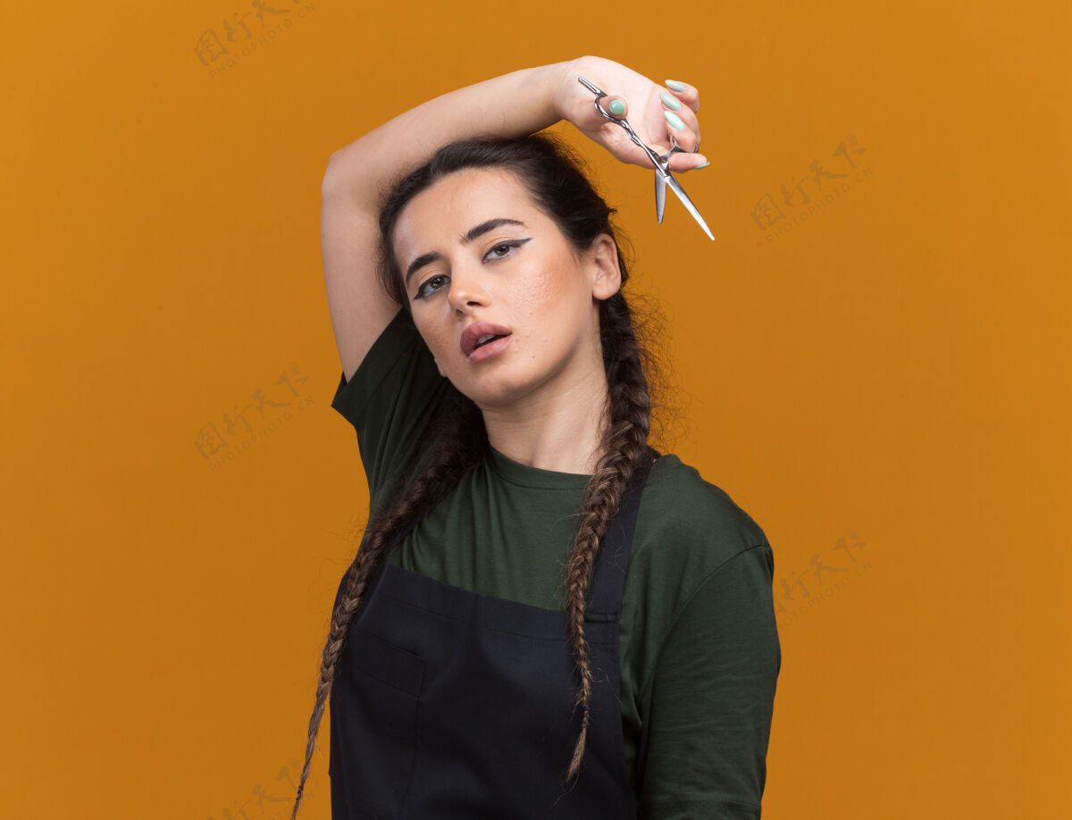 拿着印象深刻的年轻女理发师穿着制服拿着剪刀在肩膀上孤立的橙色墙上与复制空间理发师印象市民