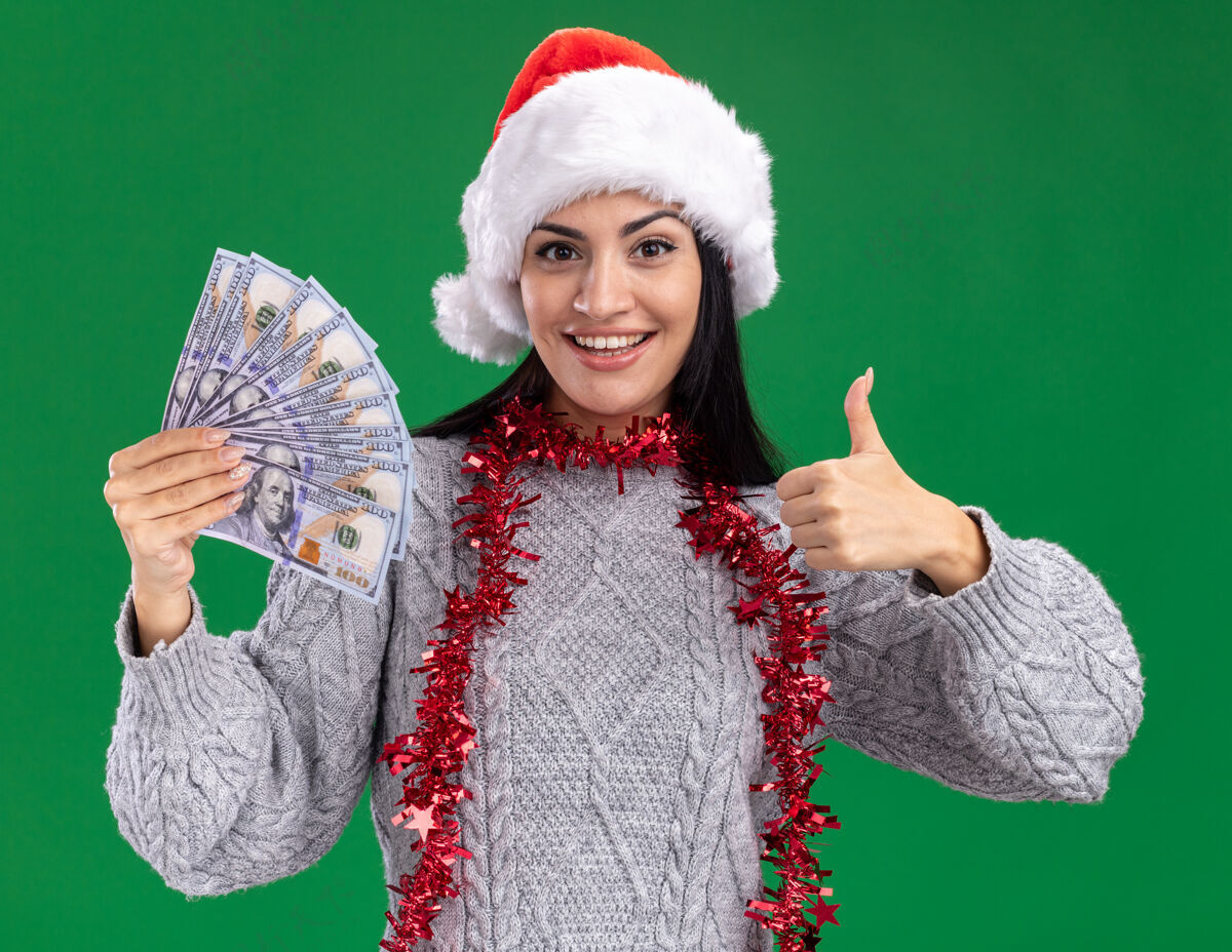周围快乐的年轻白人女孩戴着圣诞帽 脖子上戴着金箔花环 手里拿着钱 看着相机 在绿色背景上孤立地竖起大拇指圣诞节向上圣诞快乐