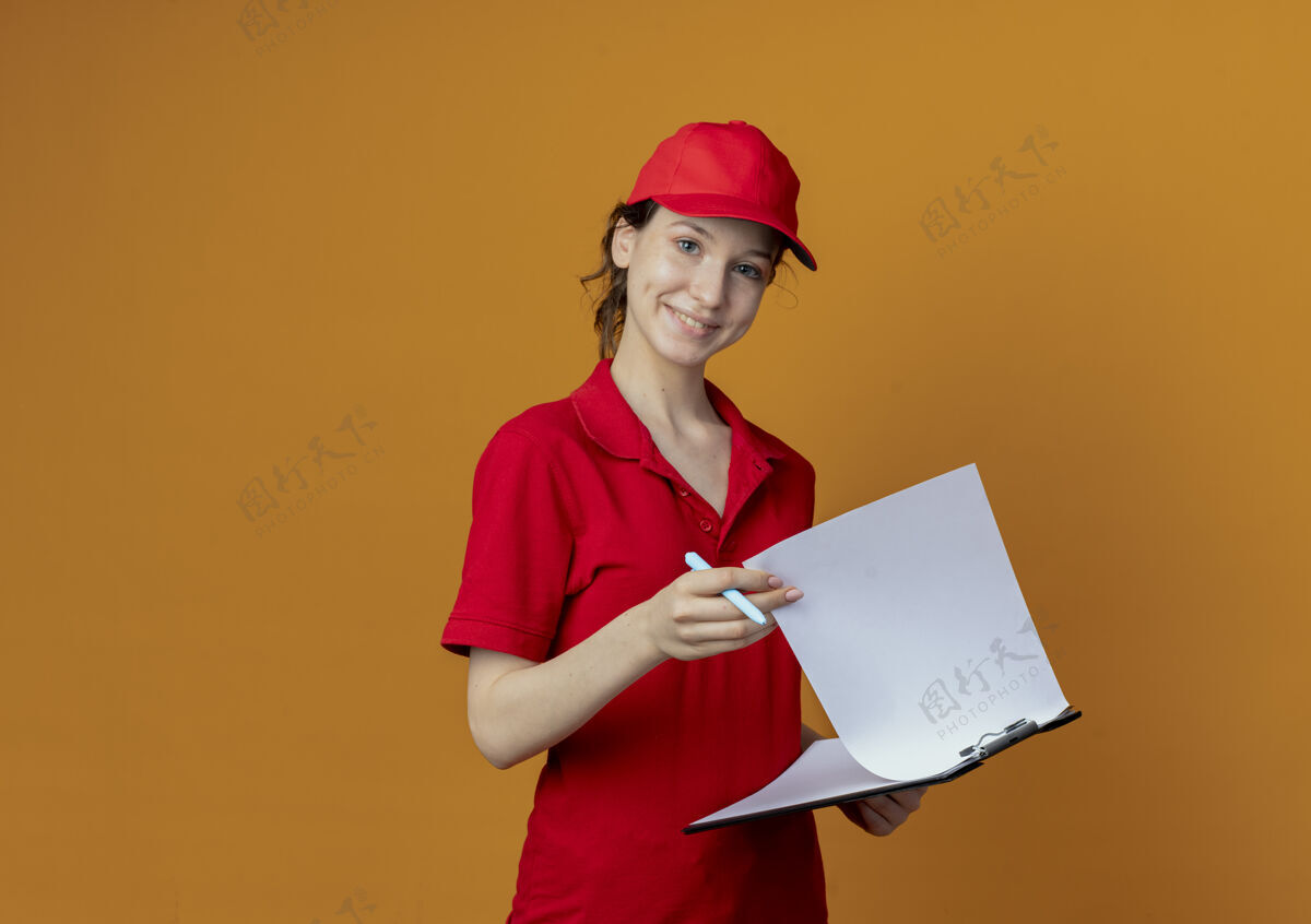 漂亮微笑着的年轻漂亮的送货女孩 穿着红色制服 戴着帽子 拿着剪贴板和笔 孤立地放在橙色背景上 还有复印空间微笑拿着帽子
