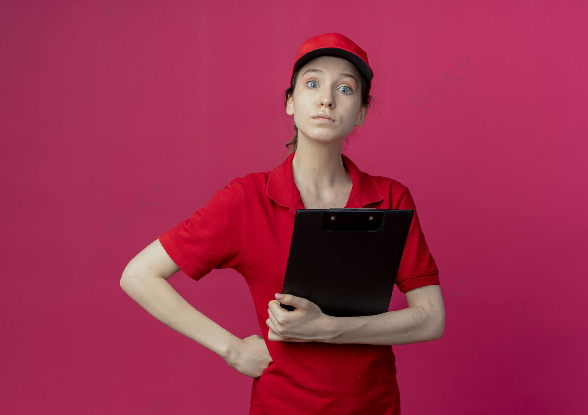 交货令人印象深刻的年轻漂亮的送货女孩 穿着红色制服 戴着帽子 拿着剪贴板 把手放在腰上 在深红色背景上 留有复印空间剪贴板帽子漂亮