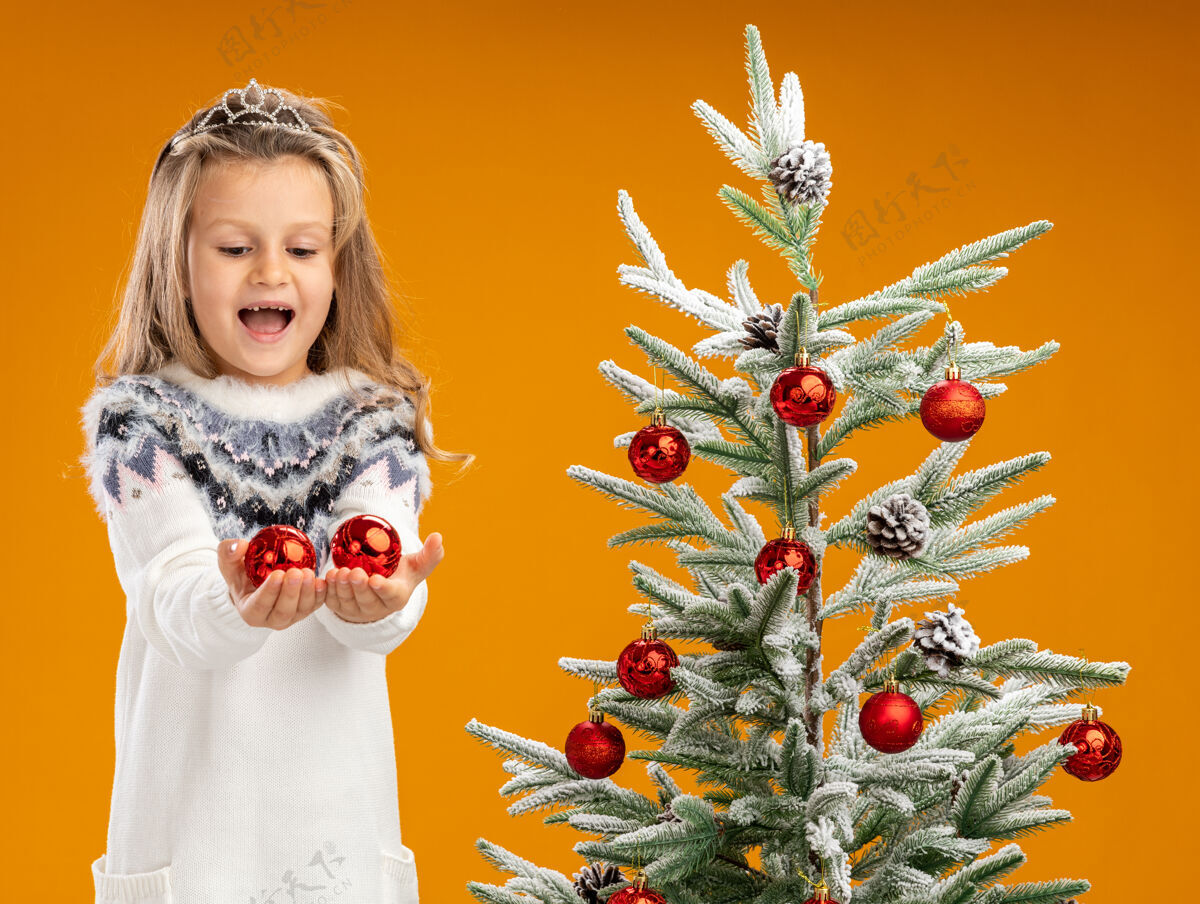 球快乐的小女孩站在圣诞树旁 戴着头饰 脖子上戴着花环 对着橙色背景的摄像机 举着圣诞球女孩橙色圣诞快乐