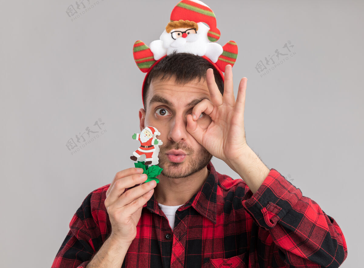 年轻印象深刻的年轻白种人戴着圣诞老人的头带拿着雪人圣诞玩具看着相机做的表情手势孤立在白色背景上举行雪人圣诞