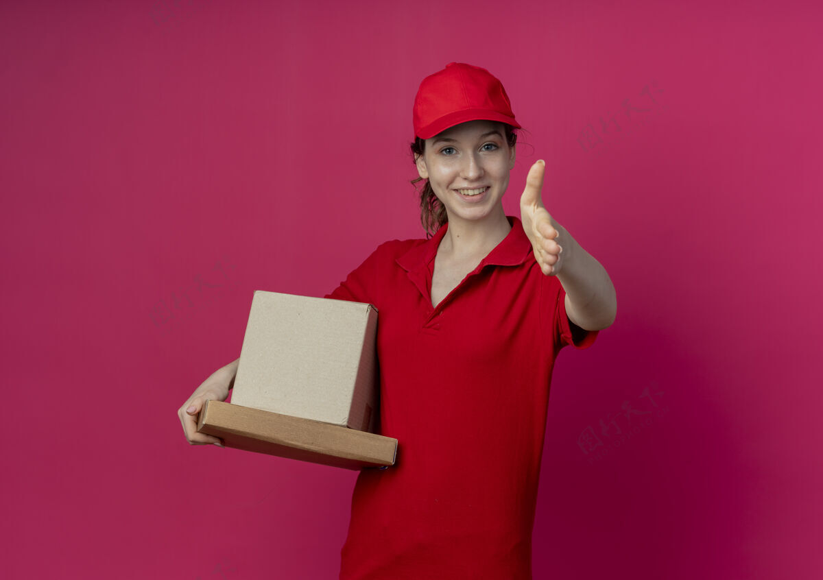 拿着微笑着的年轻漂亮的送货女孩 穿着红色制服 戴着帽子 手里拿着披萨包和纸箱 向镜头伸出手来 在深红色的背景和复印空间里 向大家打招呼送货朝向深红色