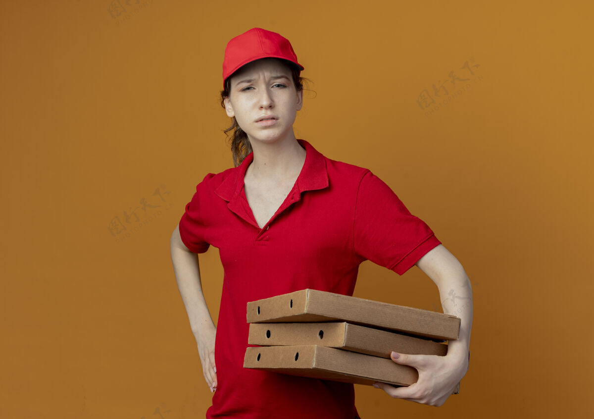 不愉快年轻漂亮的送货员 穿着红色制服 戴着帽子 手里拿着披萨包 手放在腰上 橙色背景上 留有复印空间帽子橙色拿着