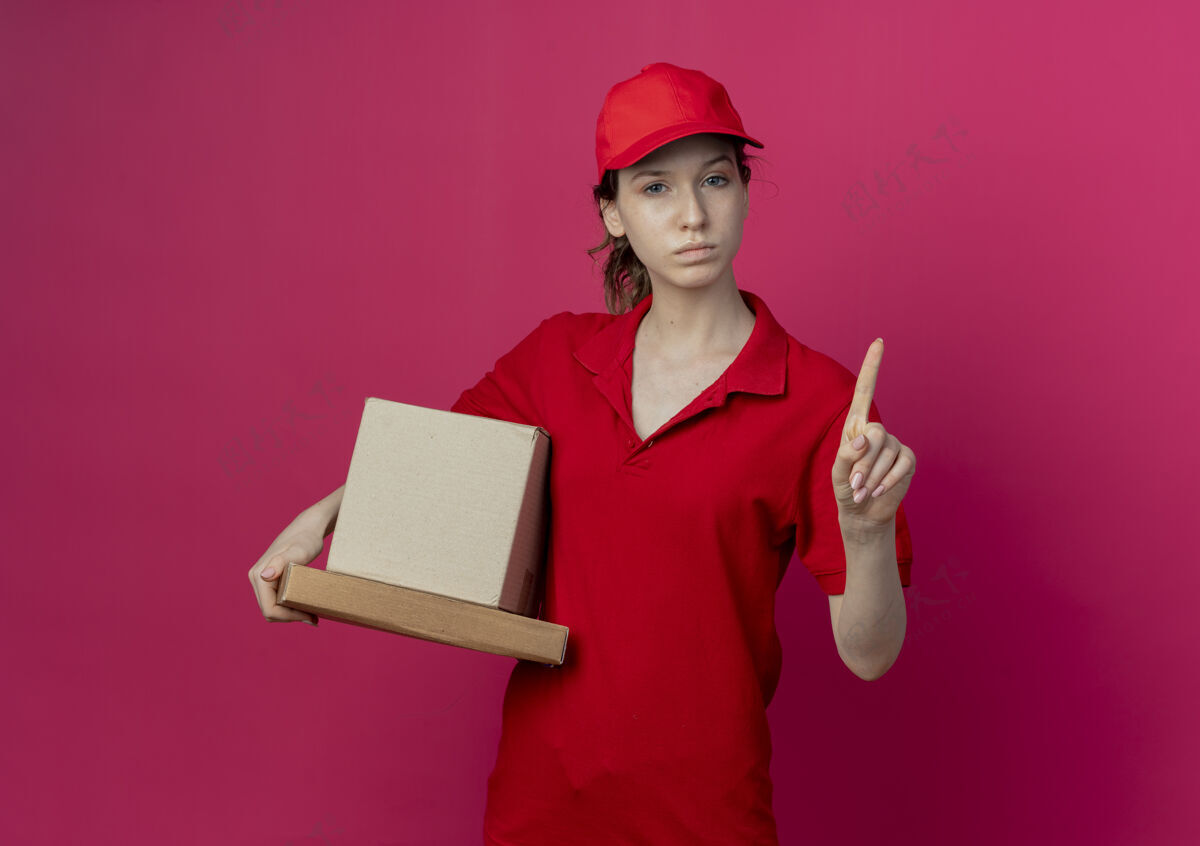 纸箱严格年轻漂亮的送货女孩穿着红色制服 戴着帽子拿着披萨包和纸箱举起手指示意没有孤立的深红色背景与复制空间严格盒子年轻