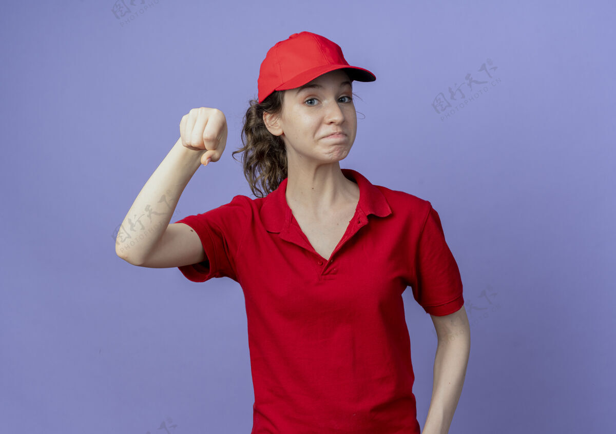交货高兴的年轻漂亮的送货女孩穿着红色制服 戴着帽子假装拿着紫色背景上的复制空间孤立的东西请年轻紫色