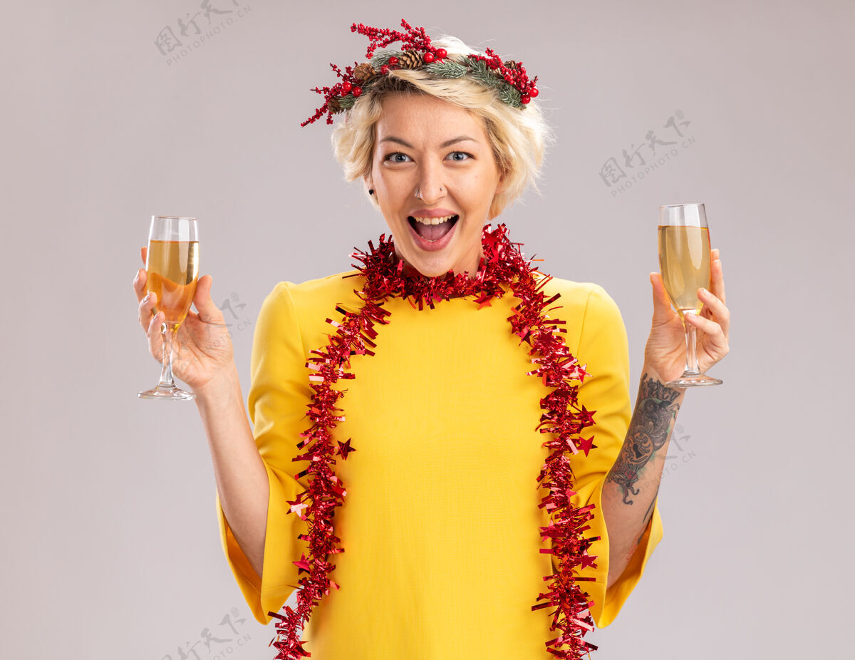 女人令人印象深刻的金发女郎戴着圣诞花环 脖子上戴着金箔花环 手里拿着两杯香槟 看着隔离在白色背景上的摄像机周围两个香槟