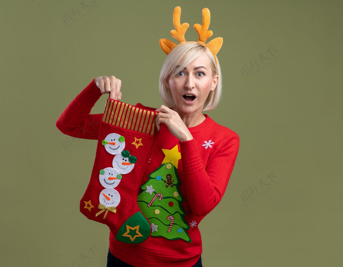 女人令人印象深刻的中年金发女人戴着圣诞驯鹿鹿角头带和圣诞毛衣拿着圣诞长袜看着橄榄绿背景上孤立的相机圣诞节举行驯鹿