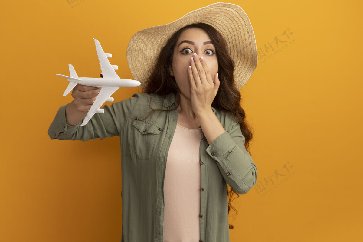 帽子害怕的年轻漂亮女孩穿着橄榄绿t恤 戴着帽子拿着玩具飞机捂着嘴 手被隔离在黄色的墙上嘴人姿势