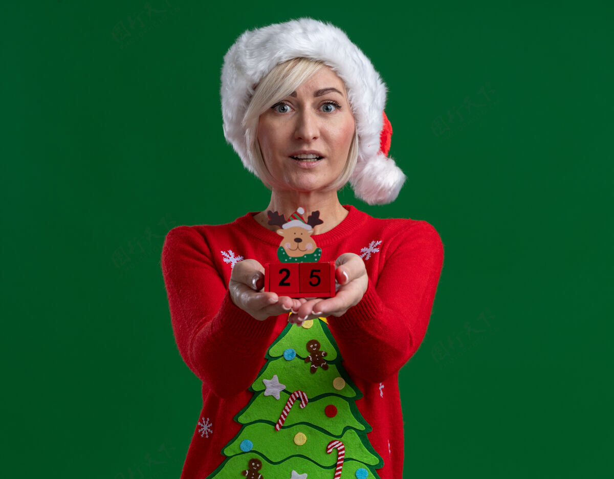 圣诞快乐印象深刻的中年金发女人戴着圣诞帽和毛衣伸展圣诞驯鹿玩具与日期对相机看着相机隔离在绿色的背景空间绿色女人