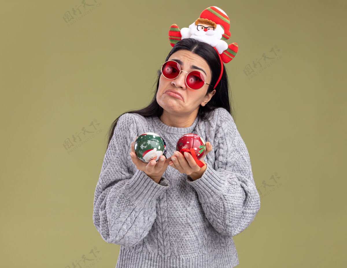 圣诞老人无知的年轻白人女孩戴着圣诞老人的头带 戴着眼镜 拿着圣诞饰品 看着橄榄绿背景上的相机女孩圣诞老人眼镜