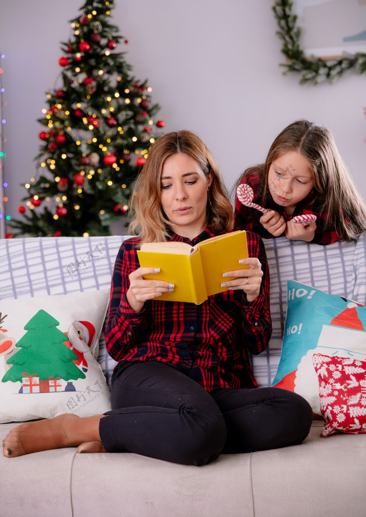 圣诞快乐女儿拿着糖果手杖 看着妈妈坐在沙发上看书 在家里享受圣诞节时光时间享受沙发