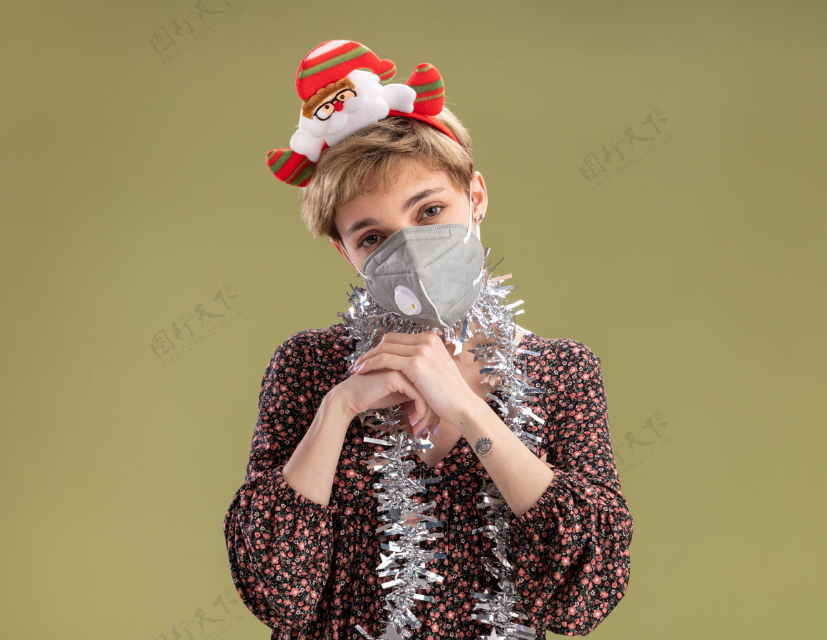 手狡猾的年轻漂亮女孩戴着圣诞老人的头带 脖子上戴着金属丝的花环 戴着防护面具 看着相机 双手在橄榄绿的背景上保持着隔离金属丝一起圣诞老人