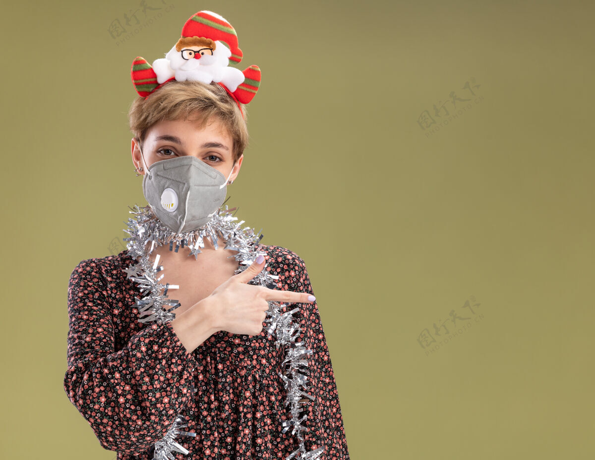 花环年轻漂亮的女孩戴着圣诞老人的头带 脖子上戴着金属丝的花环 戴着防护面具 看着摄像机指着橄榄绿背景上孤立的一边空间面具脖子