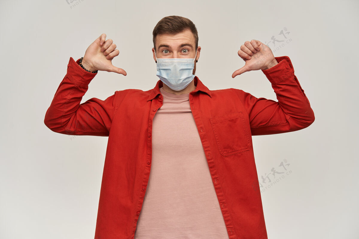 年轻身穿红衫 脸上戴着防病毒口罩 满脸胡须的快乐年轻人用两只大拇指指着自己的白墙面具健康衬衫