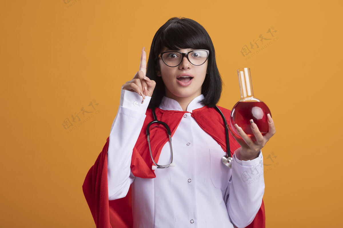 听诊器令人印象深刻的年轻超级英雄女孩戴听诊器与医疗袍和斗篷与眼镜举行化学玻璃瓶装满红色液体印象斗篷化学