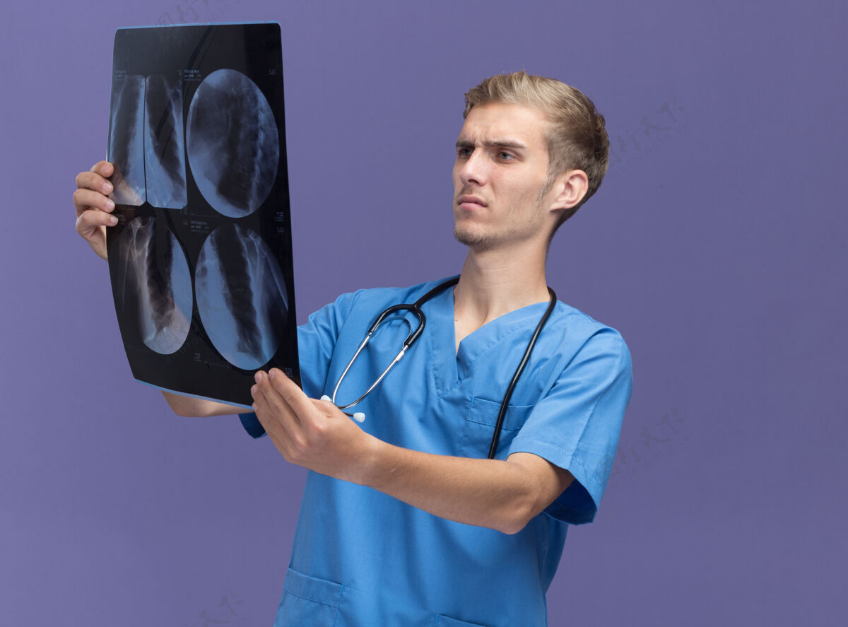 站立想着年轻的男医生穿着医生制服拿着听诊器看隔离在蓝色墙上的x光片表情穿着感觉