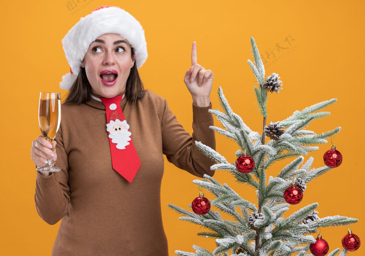 香槟给人印象深刻的年轻漂亮女孩戴着圣诞帽打着领带站在圣诞树旁拿着一杯香槟点在橙色背景上孤立起来帽子向上玻璃杯