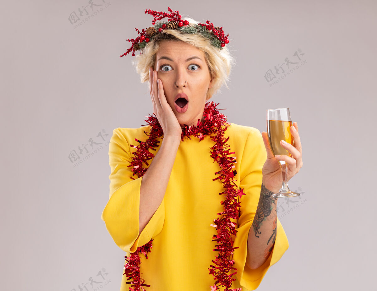 关心关心的金发女郎戴着圣诞花环 脖子上戴着金箔花环 手里拿着一杯香槟 看着镜头 手放在脸上 在白色背景下与世隔绝金发头年轻