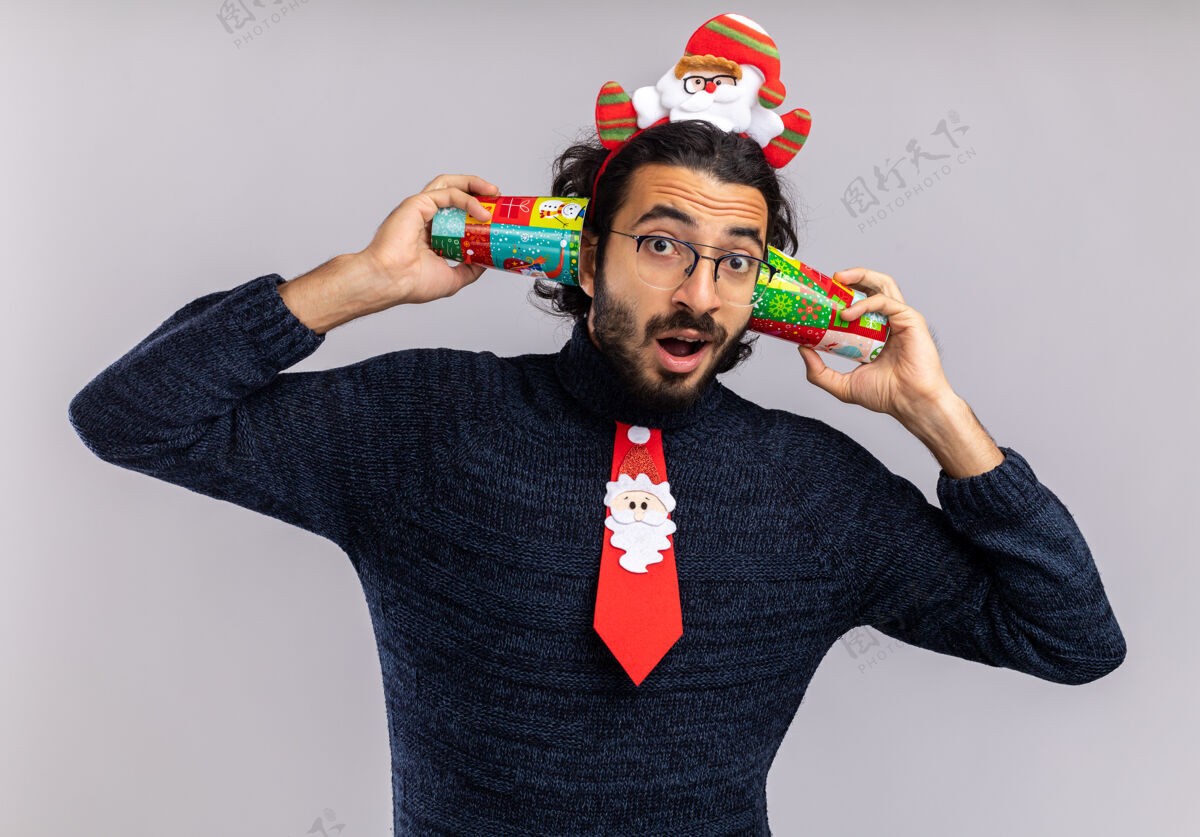 小伙子印象深刻的年轻帅哥戴着圣诞领带和发箍把圣诞杯放在耳朵上隔离在白色背景上圣诞快乐领带年轻