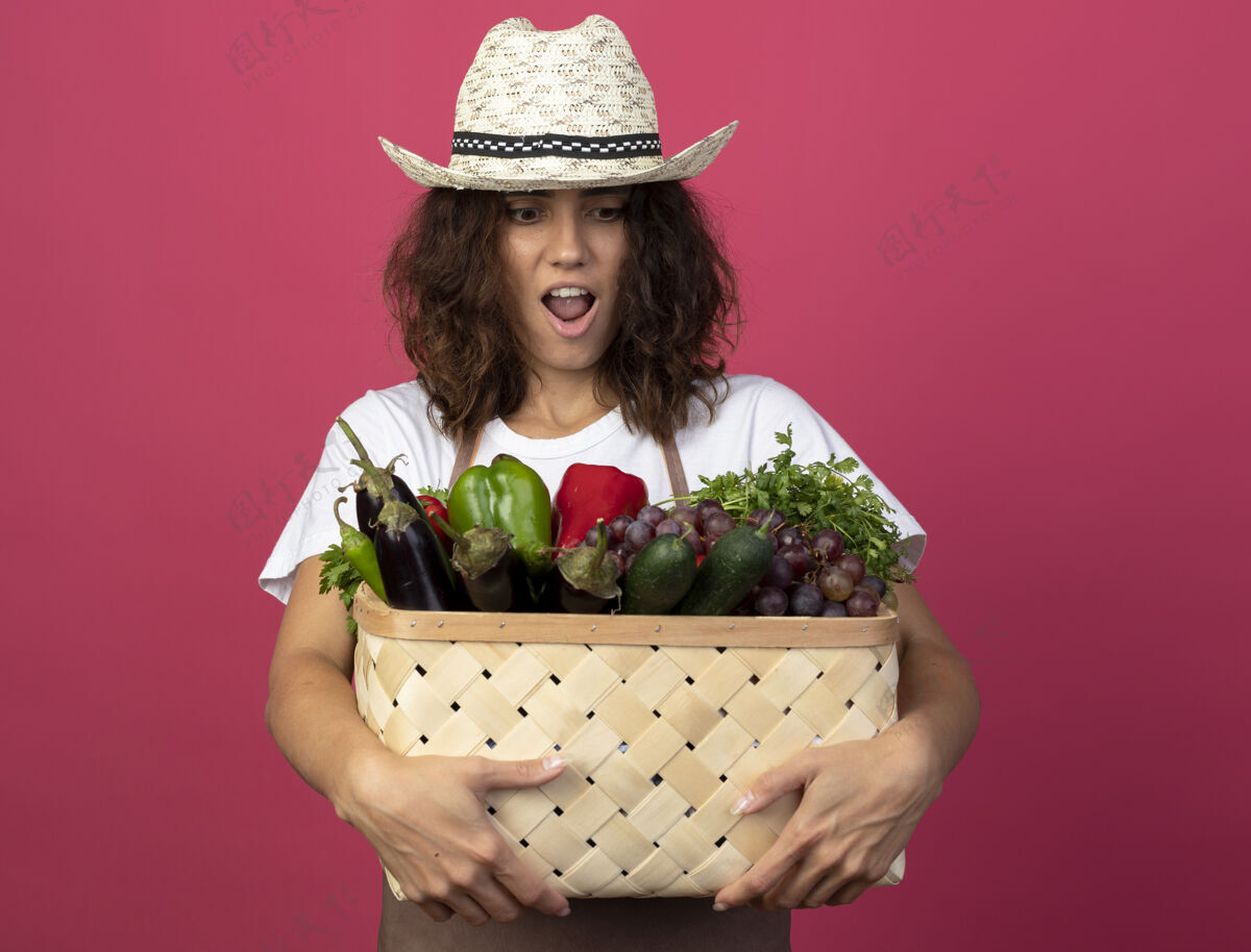 成人穿着制服的年轻女园丁惊讶地戴着园艺帽 拿着菜篮看着制服穿举行