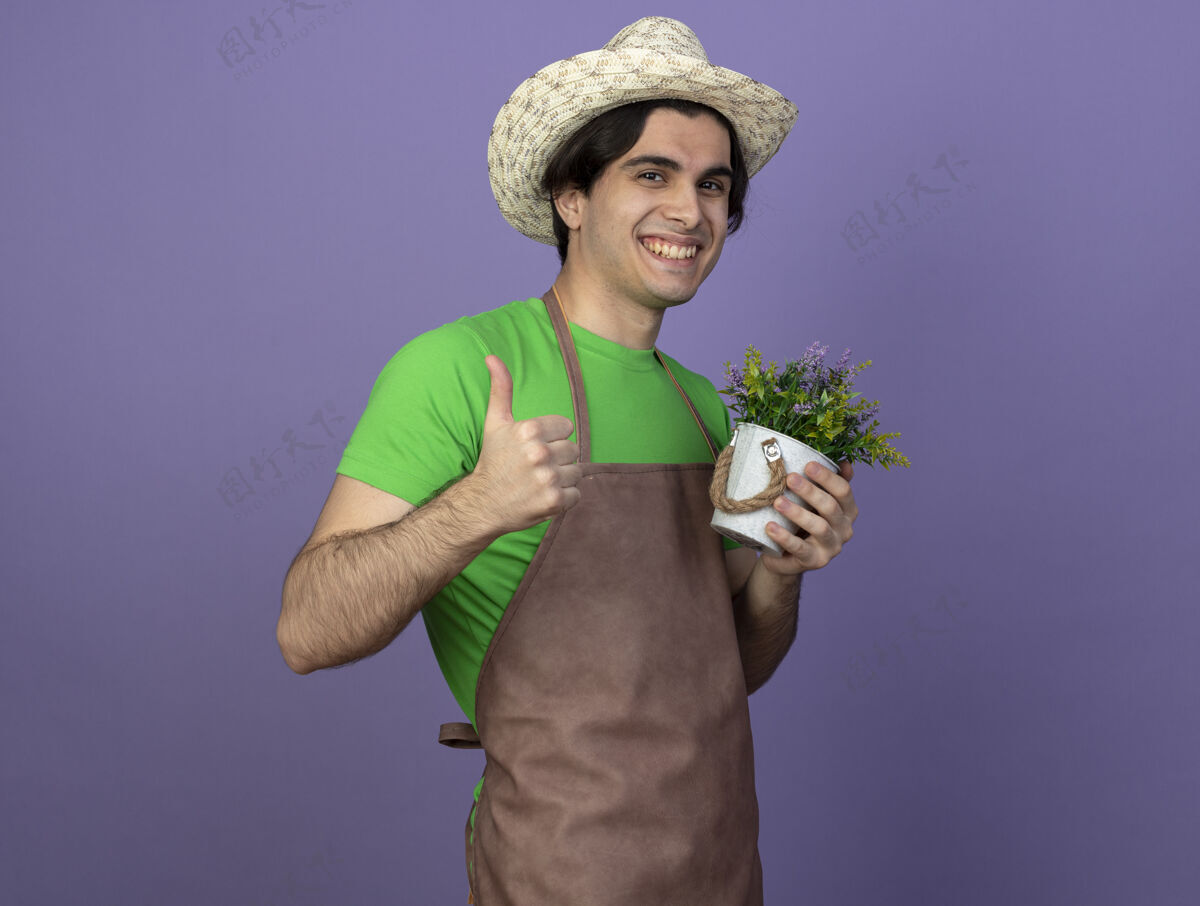 花盆微笑着的年轻男园丁穿着制服 戴着园艺帽 手里拿着花盆里的花 竖起大拇指向上紫色微笑