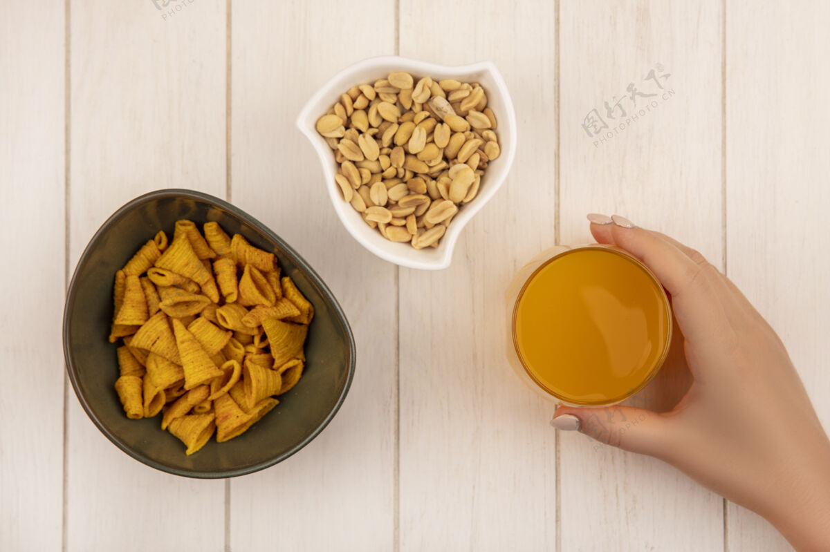 橙子俯视图：米色木桌上的白色碗上 一只手拿着一杯橙汁和一碗锥形玉米小吃和松子木头薯片形状