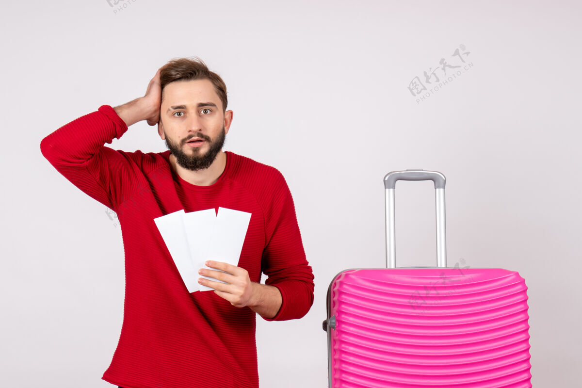 微笑正面图：年轻男性 带着粉红色的包 手持机票 在白色的墙上航行 飞行 彩色旅行 度假照片 情感风景年轻的男性飞行