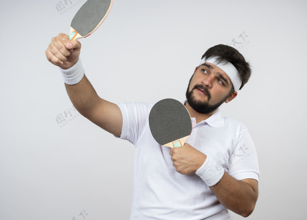手持高兴的年轻人看着一边戴着头带和手环拿着乒乓球拍隔离在白色的墙上请戴球拍头带