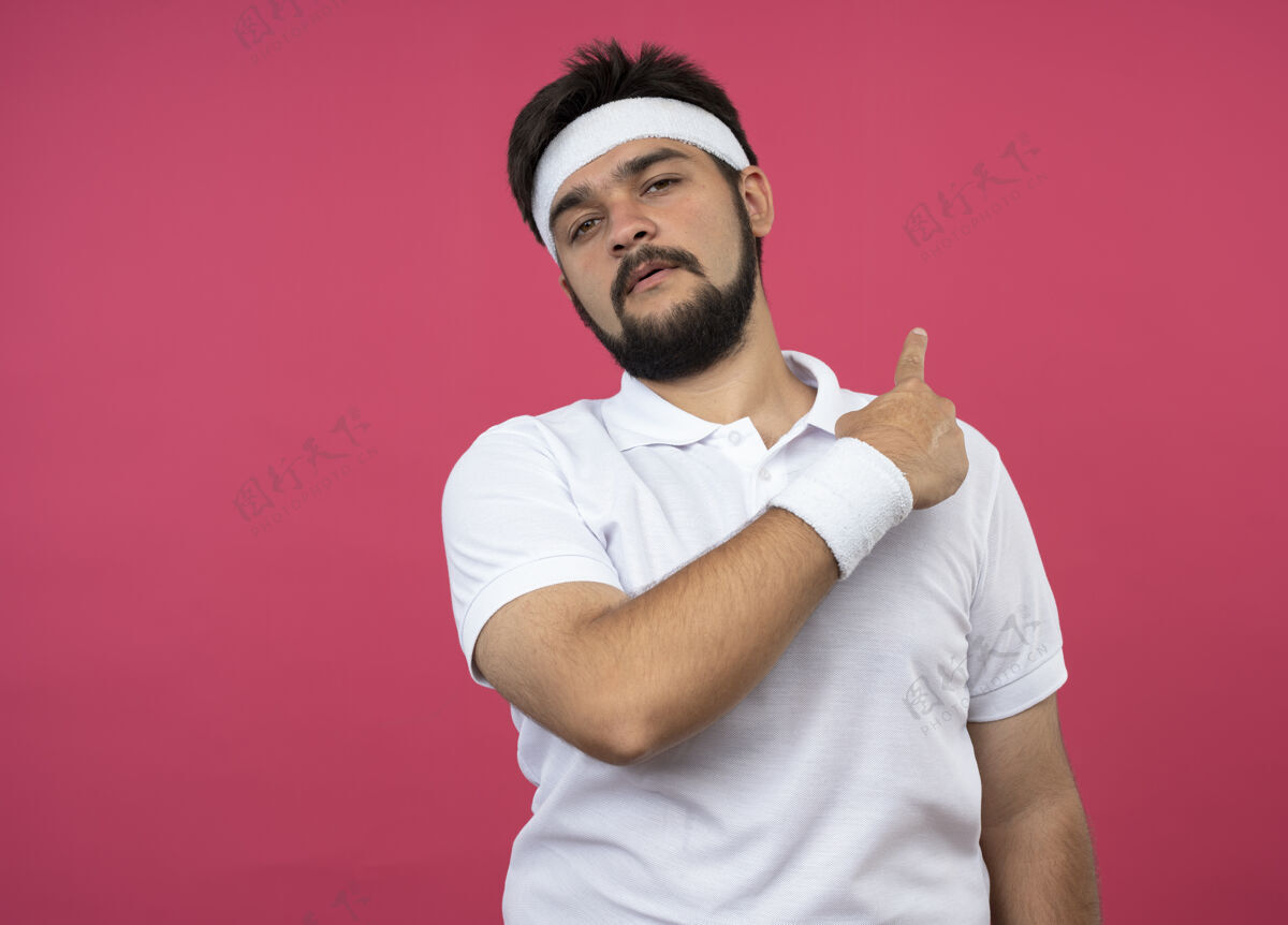年轻戴着头带和腕带的未被释放的年轻运动型男子指向后面 隔离在粉色墙壁上 留有复制空间男人头带运动
