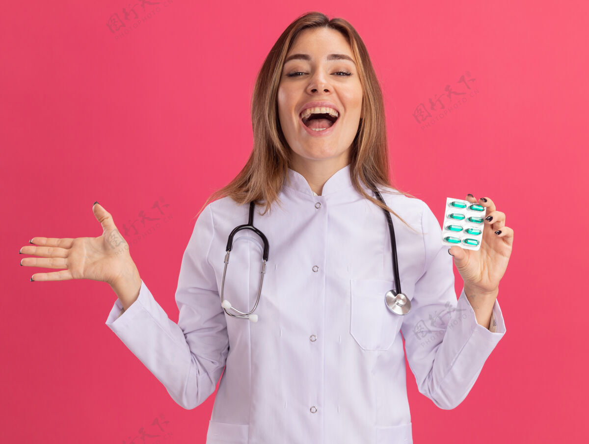 抱着兴奋的年轻女医生 穿着医用长袍 手持听诊器 手被隔离在粉红色的墙上 手里拿着药片传播药丸穿
