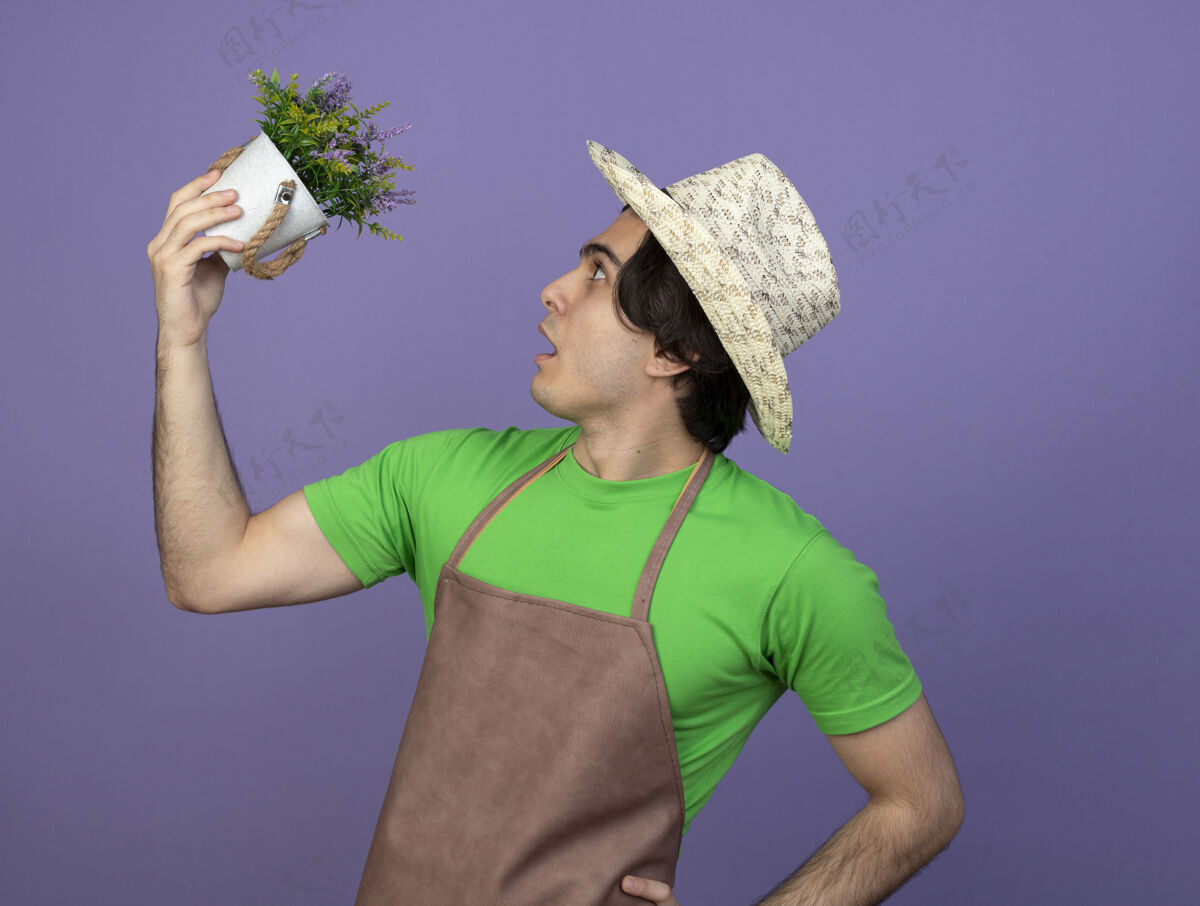 帽子惊讶地站在侧视图年轻的男园丁穿着制服戴着园艺帽抬起头看着花盆里的花把手放在屁股上孤立的紫色花园穿园艺