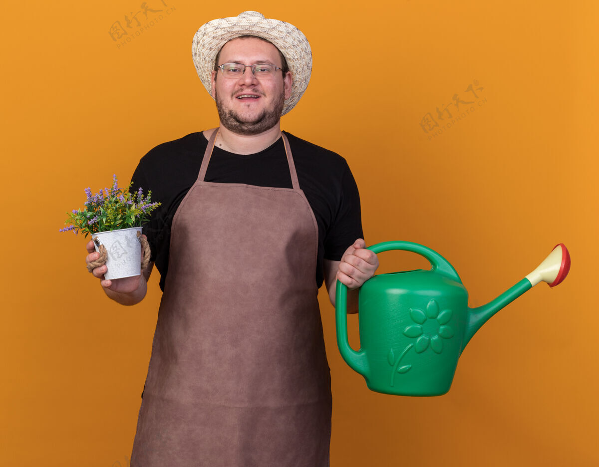 年轻高兴的年轻男园丁戴着园艺帽和手套拿着浇水罐 花盆里的花被隔离在橙色的墙上穿请帽子