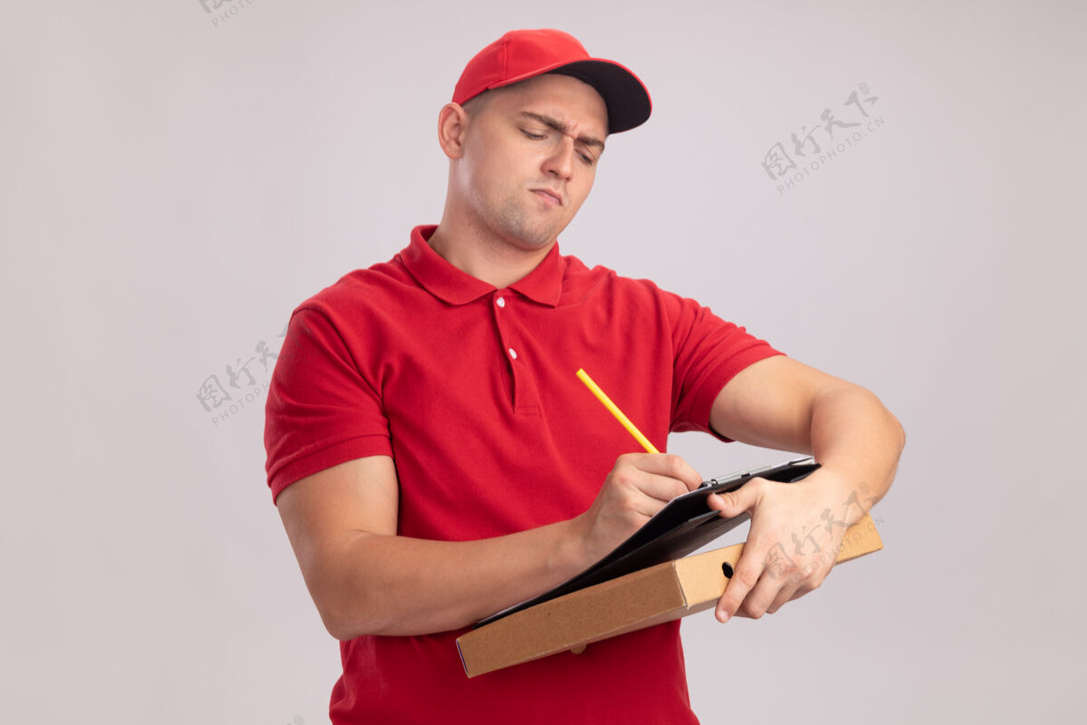 写字自信的年轻送货员穿着制服 戴着帽子 拿着披萨盒 在白墙上的剪贴板上写着什么自信男人什么的
