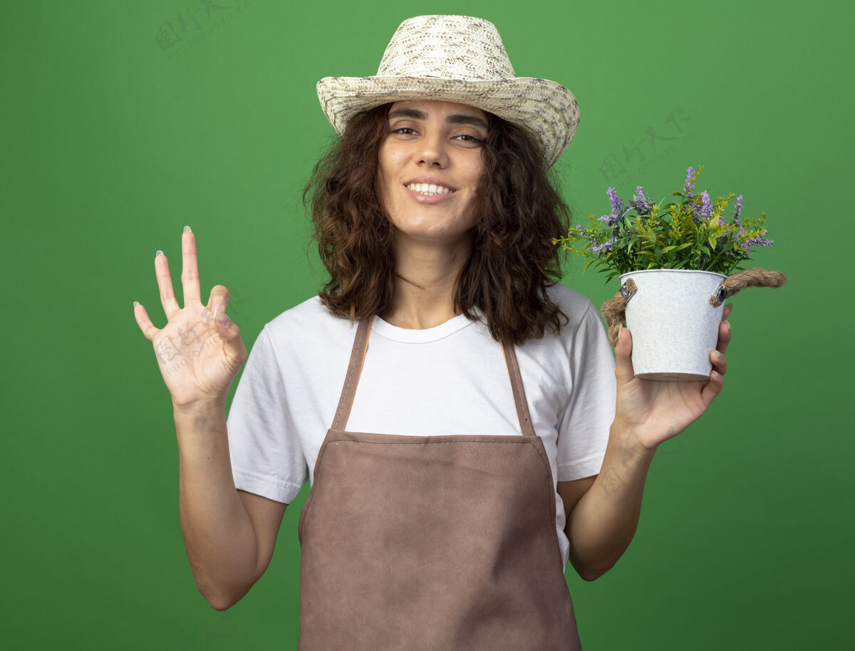 花盆身着制服 面带微笑的年轻女园丁戴着园艺帽 手里拿着花盆里的花 在绿茵场上表现出良好的姿态女性举行手势
