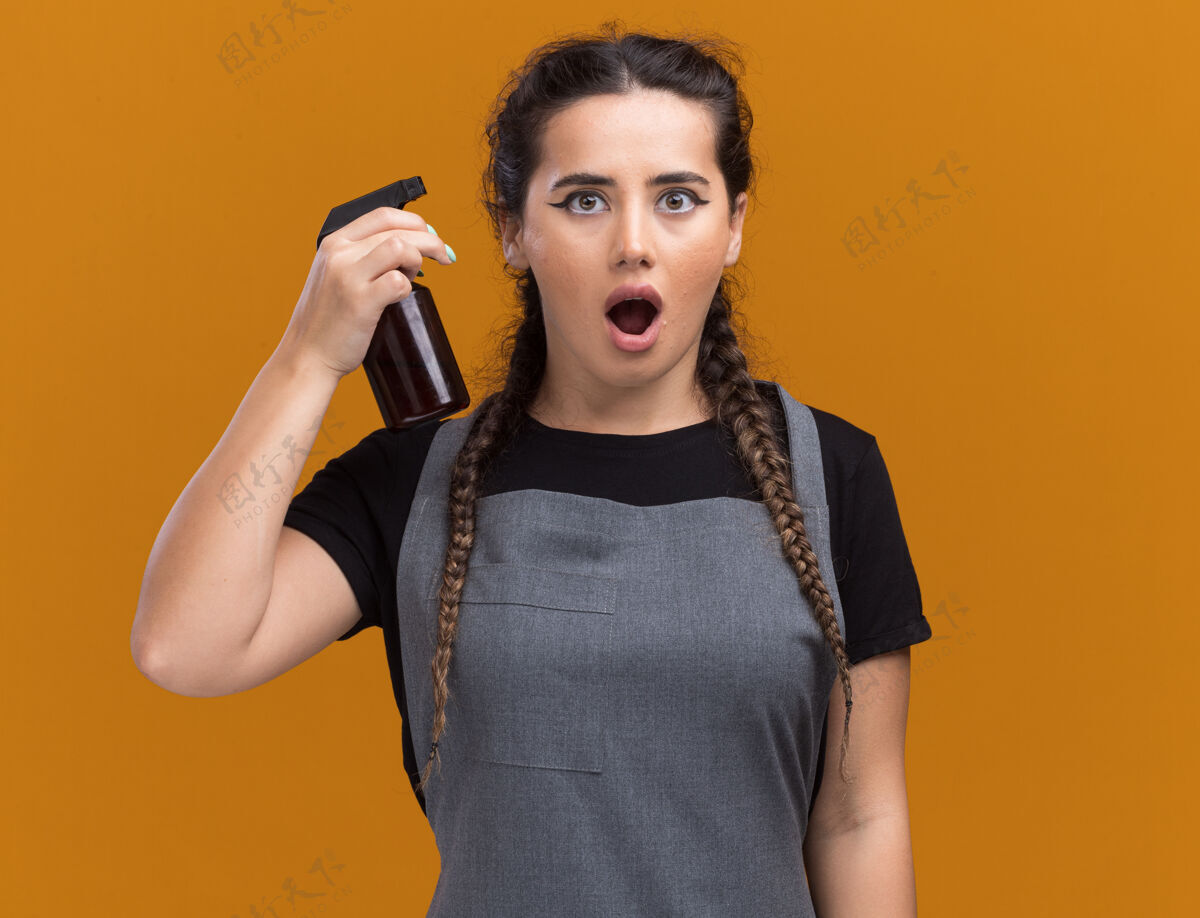 奶瓶惊讶的年轻女理发师穿着制服拿着喷雾瓶隔离在橙色的墙上拿着年轻制服