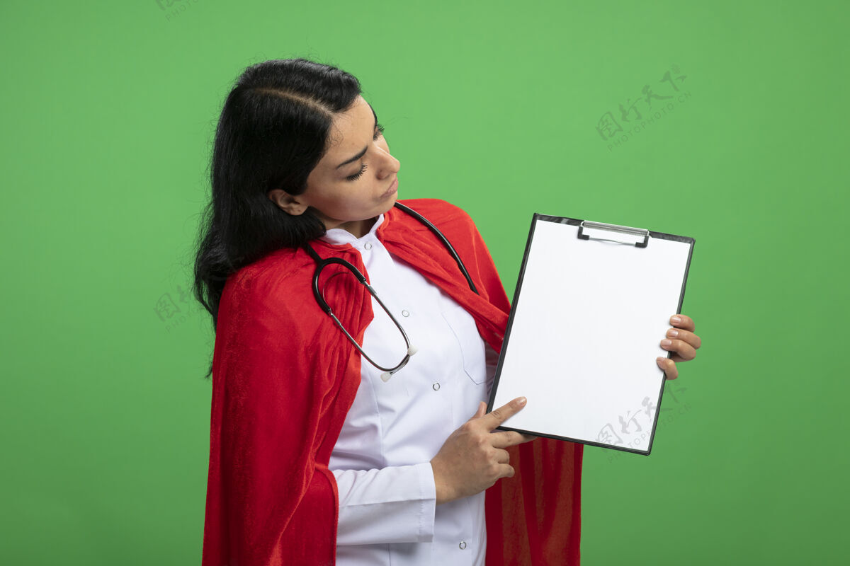 长袍沉思的年轻超级英雄女孩穿着医用长袍 手持听诊器 看着绿色的剪贴板穿举行医疗