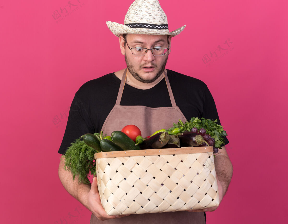 花园年轻的男园丁惊讶地戴着园艺帽 手里拿着一个孤立在粉红色墙上的菜篮子蔬菜园艺帽子