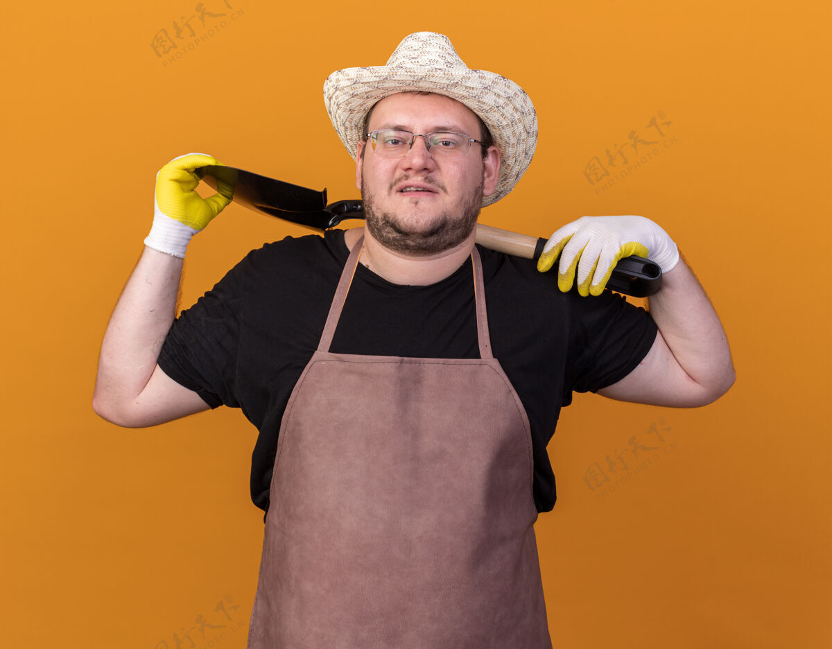 信心自信的年轻男园丁戴着园艺帽和手套 肩上扛着铁锹 隔离在橙色的墙上花园手套黑桃