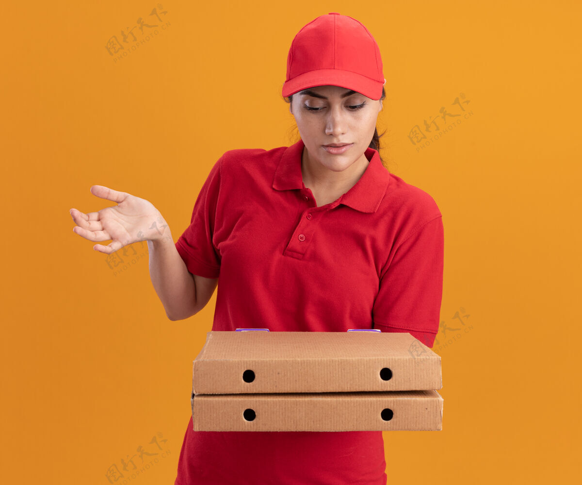 帽子困惑的年轻送货女孩穿着制服 戴着帽子 看着披萨盒摊开手孤立在橙色墙上迷糊盒子年轻人