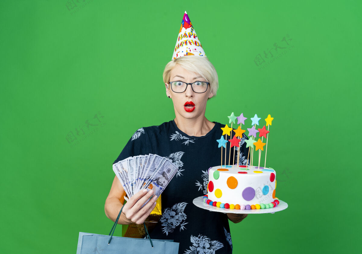 感情年轻的金发派对女郎戴着眼镜 戴着生日帽 手里拿着生日蛋糕 手里拿着星星的钱礼盒和纸袋 看着前面的绿色墙壁上孤立的复制空间生日举行立场
